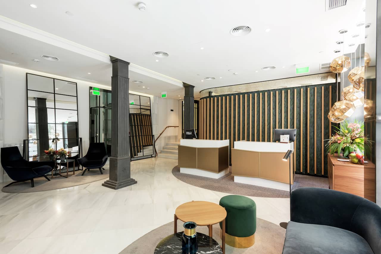 A hotel lobby with a marble floor, a curved wood-and-green reception desk, dark armchairs and a hanging gold light fixture.