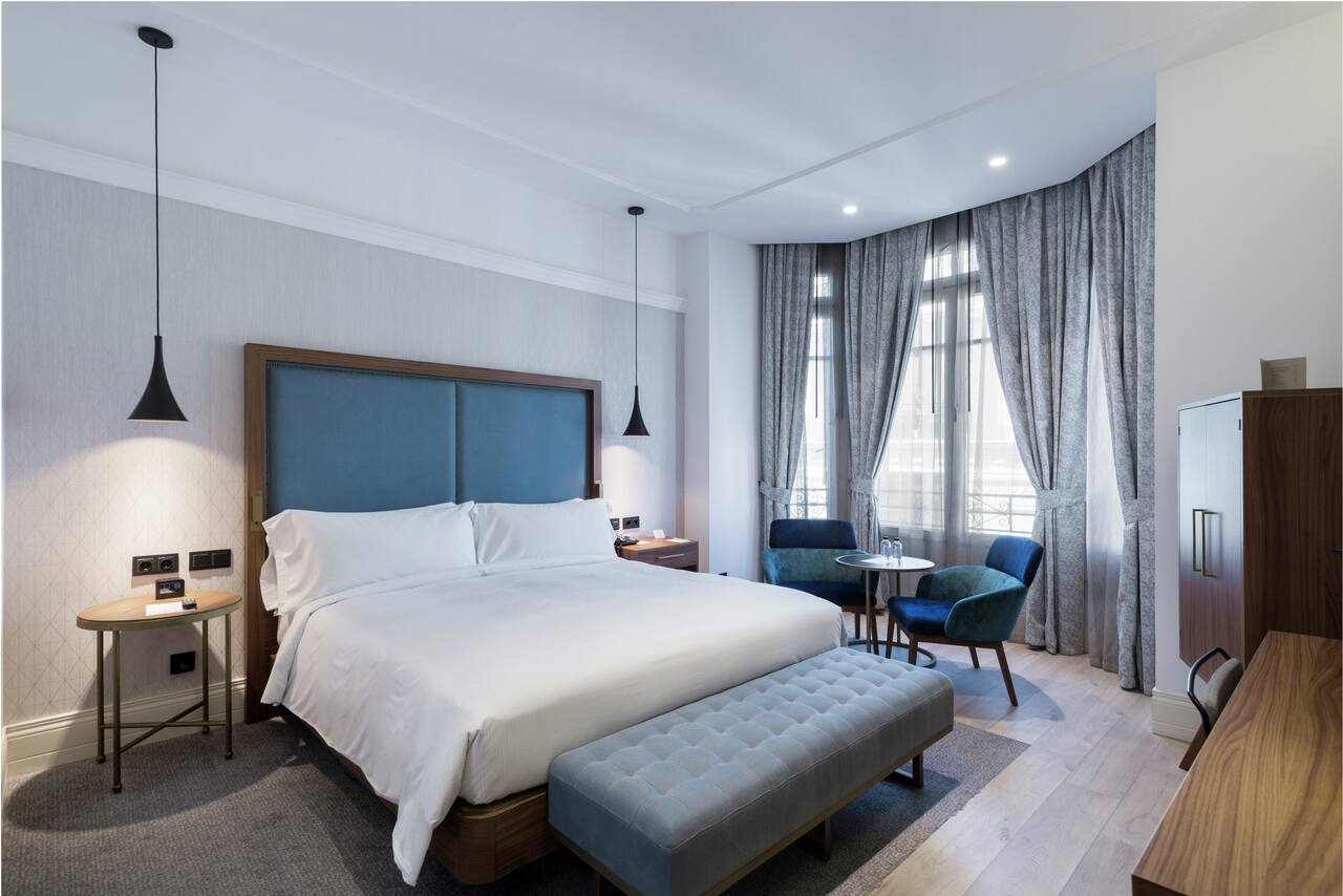 A hotel room with a blue upholstered headboard, white bedding, a grey tufted bench and tall curtained windows with a wrought-iron balcony outside.