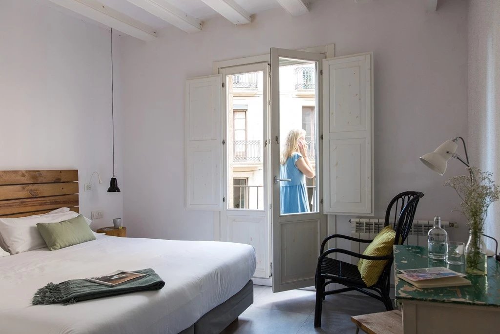Guest room at Eco Boutique Hostal Grau with a wooden headboard, white bedding, a beamed ceiling and a balcony door onto the street