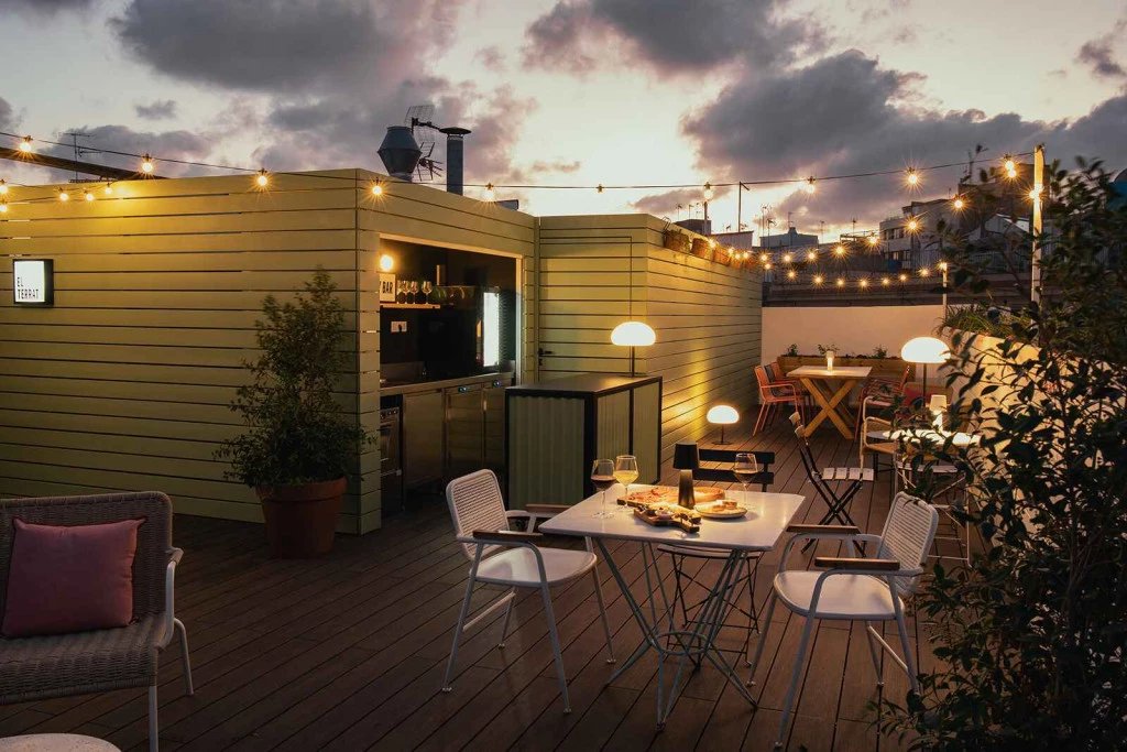 Rooftop terrace at Eco Boutique Hostal Grau at dusk, with string lights, wooden decking, plants and a small bar kiosk