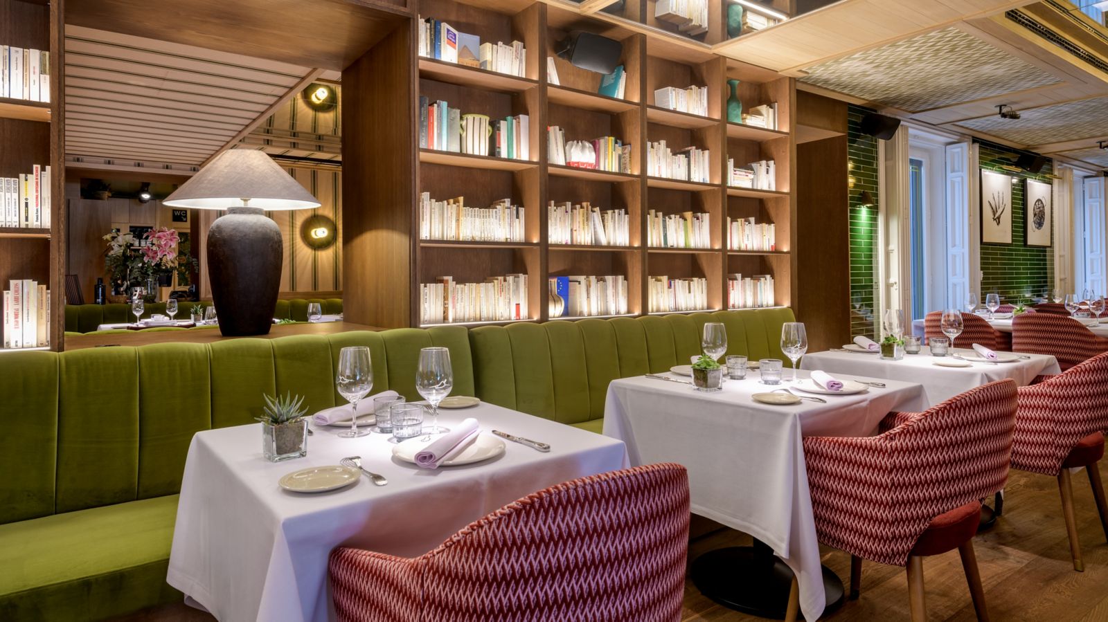 A dining corner backed by a floor-to-ceiling bookshelf wall, with set tables, green velvet banquettes, patterned armchairs and a large urn lamp.