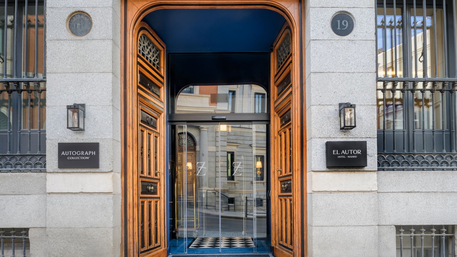 The hotel entrance at No 19: tall carved wooden doors open onto a blue vestibule with a chequered mat, flanked by lanterns and plaques reading El Autor Hotel Madrid and Autograph Collection.