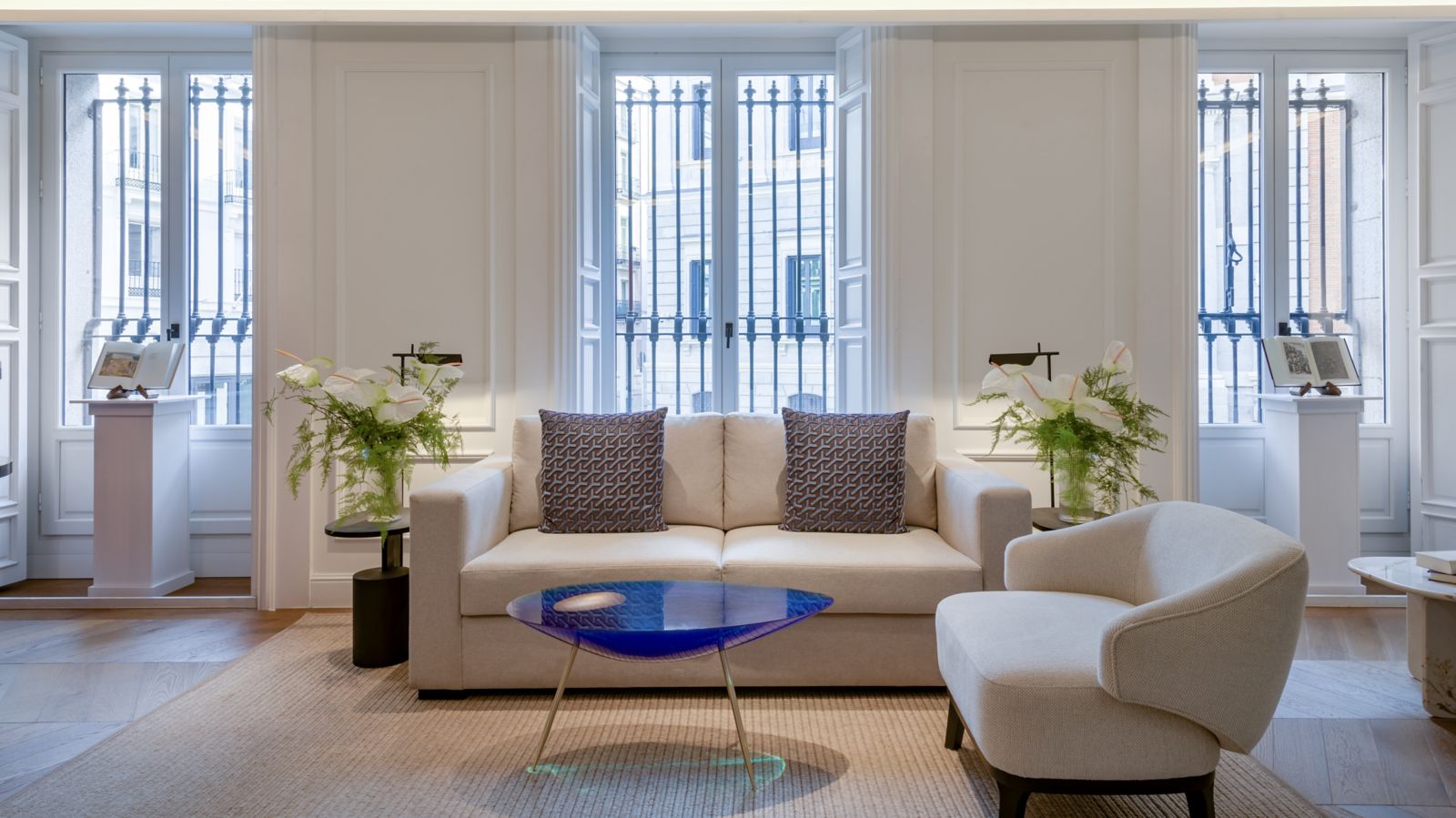 A white-panelled salon with a cream sofa and armchair around a cobalt-blue glass table, tall shuttered windows with iron grilles, and open books displayed on pedestals.