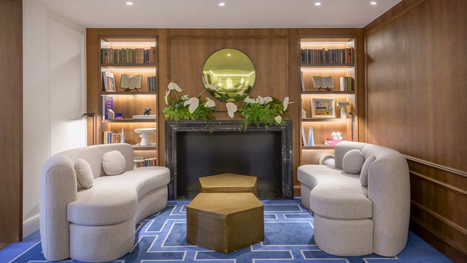 A wood-panelled library lounge: two curved boucle sofas around brass hexagonal tables on a blue geometric rug, before a black marble fireplace between lit bookshelves.