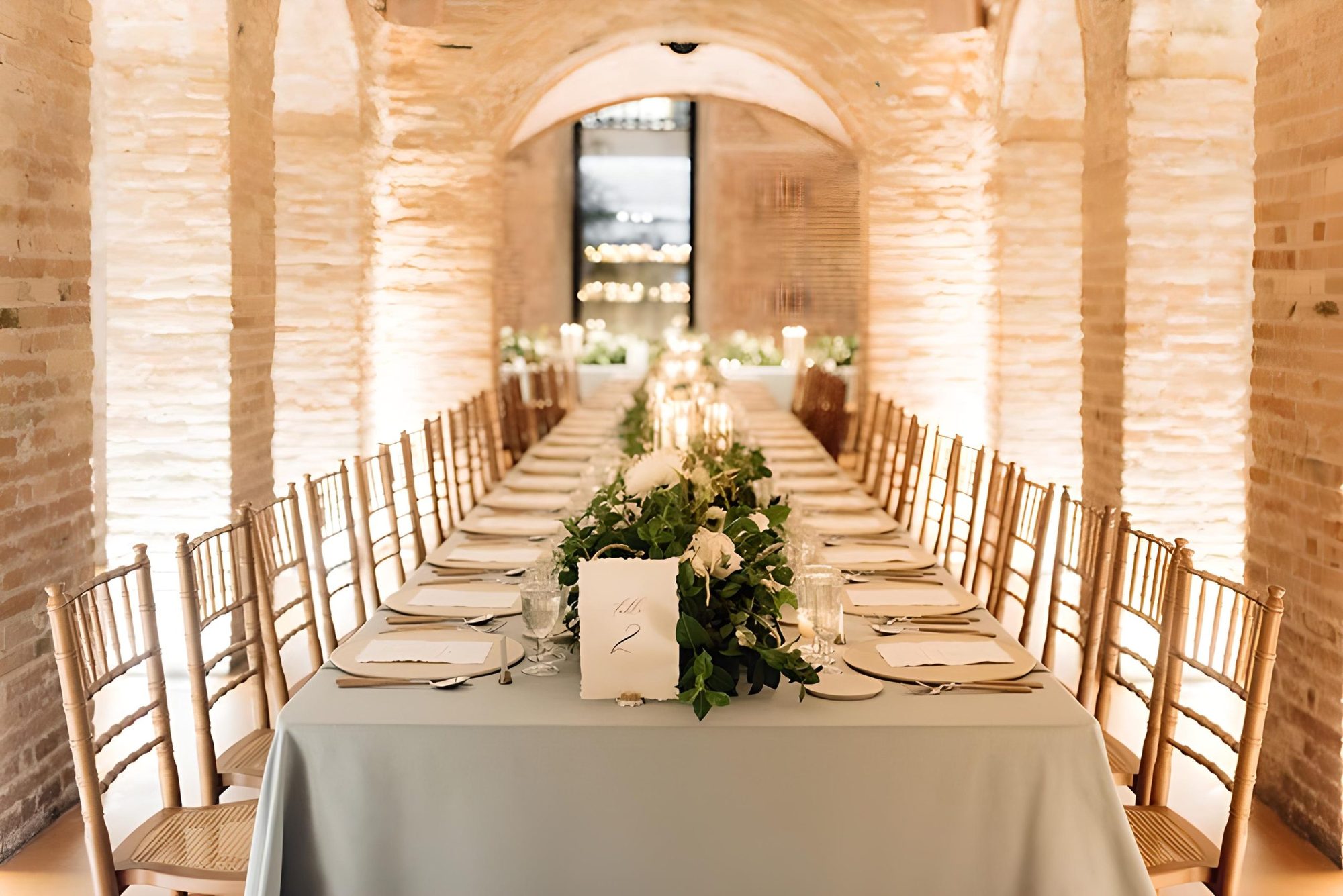 Long formal dining table set for an event under vaulted exposed-brick arches at El Palauet, with candlelight and greenery runners