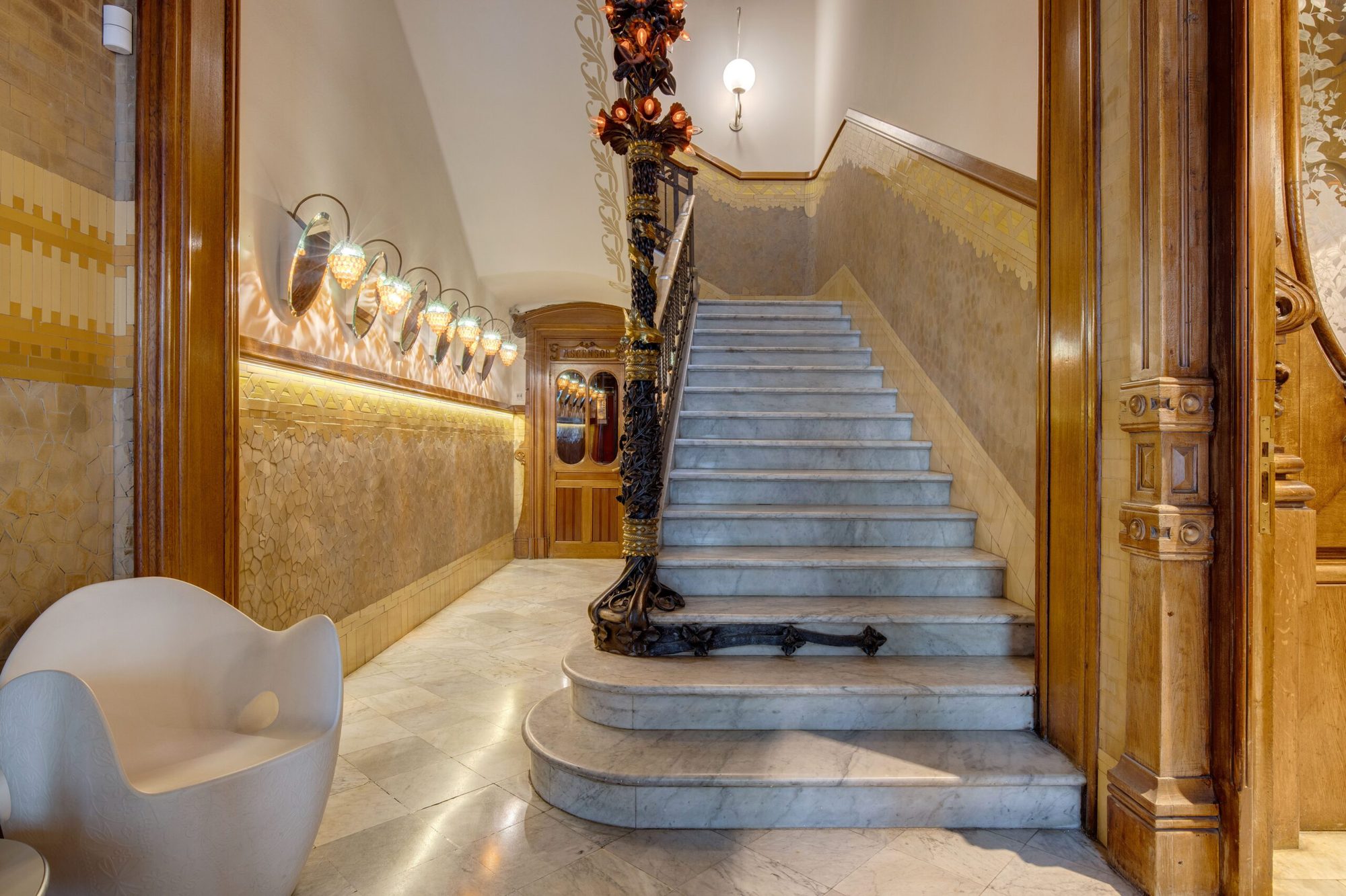 Original modernist marble staircase at El Palauet with an ornate wrought-iron banister, mosaic wall panels, and carved wood doorway
