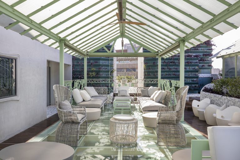 Glass-roofed rooftop conservatory lounge at El Palauet with green ironwork, wicker sofas, and city rooftops visible through the panes