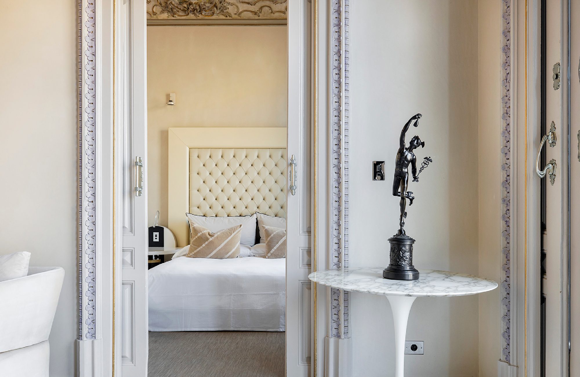 View through open double doors into a suite at El Palauet, with a tufted cream headboard, striped pillows, and a painted ceiling frieze visible above the bed
