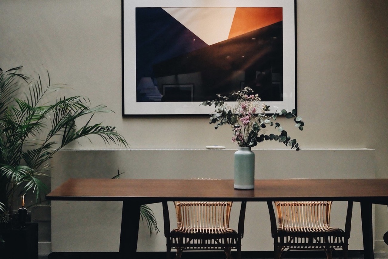 Restaurant dining table with rattan chairs, a framed abstract print and a vase of dried and fresh flowers on the tabletop.