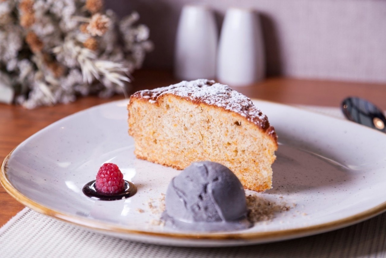 Slice of cake dusted with powdered sugar on a white plate, served with a scoop of dark sorbet and a raspberry, at FloraFina restaurant.