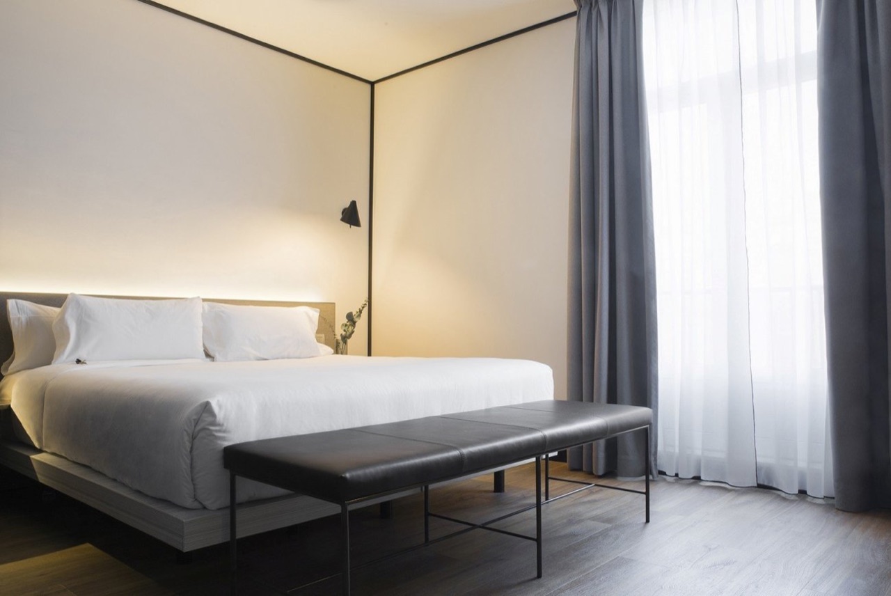 Minimalist hotel room with a white-made bed, dark headboard, leather bench and floor-to-ceiling grey curtains beside a window.