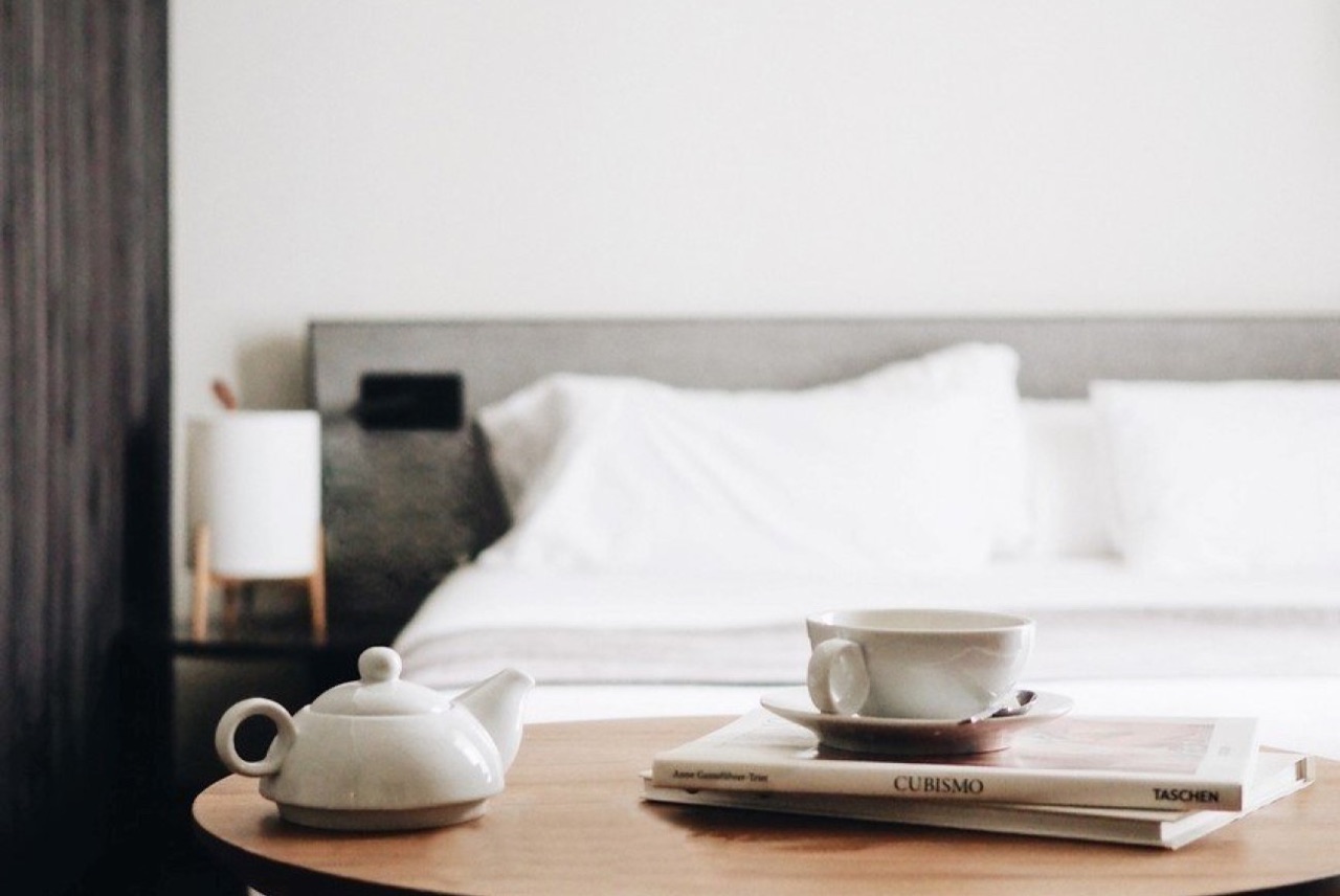 Round wooden bedside table in a hotel room with a teapot, teacup and saucer and two books, with a white-made bed blurred in the background.