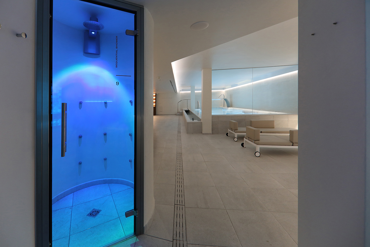 Coco Spa corridor at Es Princep with a sequential shower cabin and the indoor pool beyond