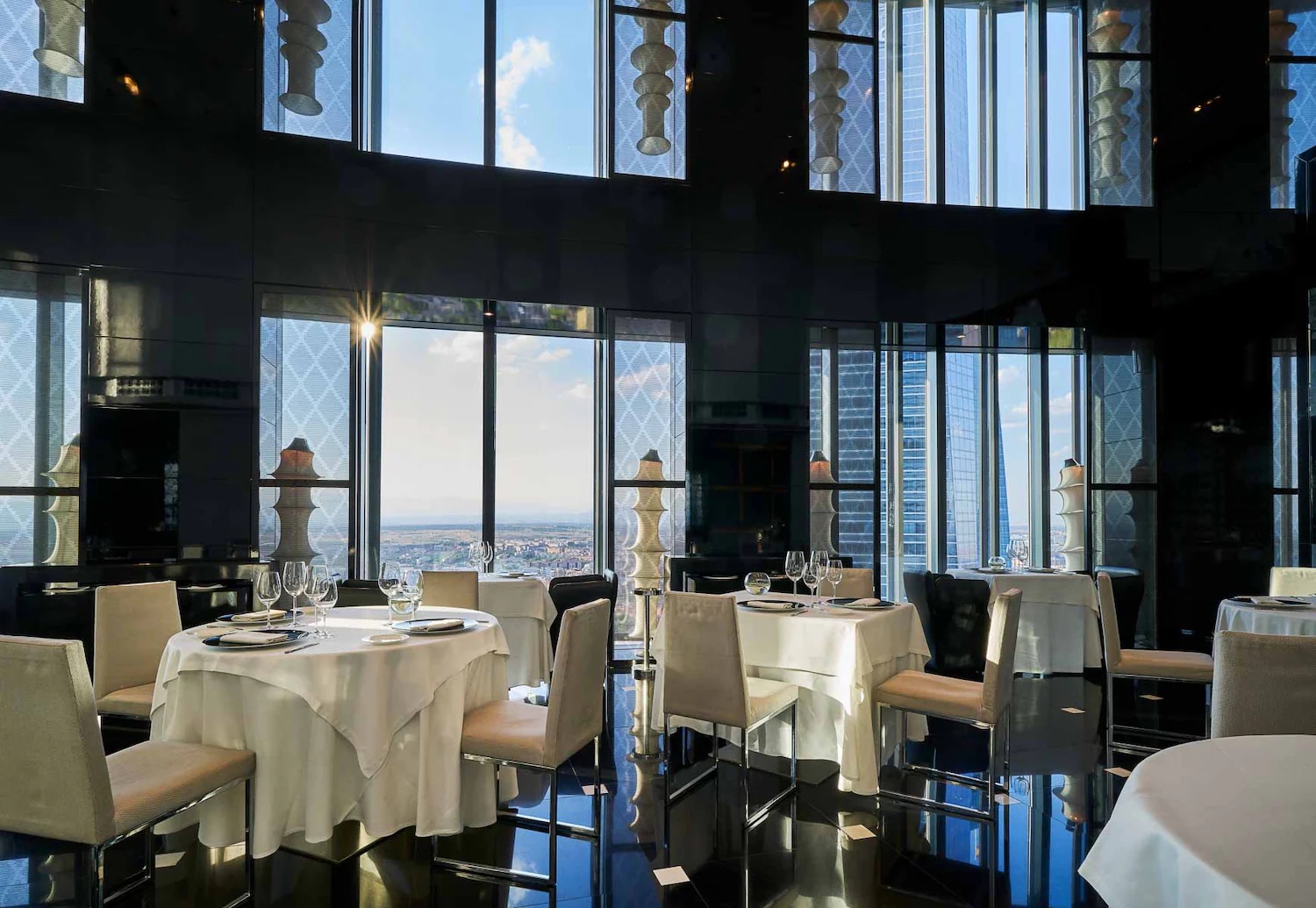 A restaurant dining room with round tables in white cloths, cream chairs and a black glossy floor, tall windows looking onto neighbouring skyscrapers and the plain beyond.