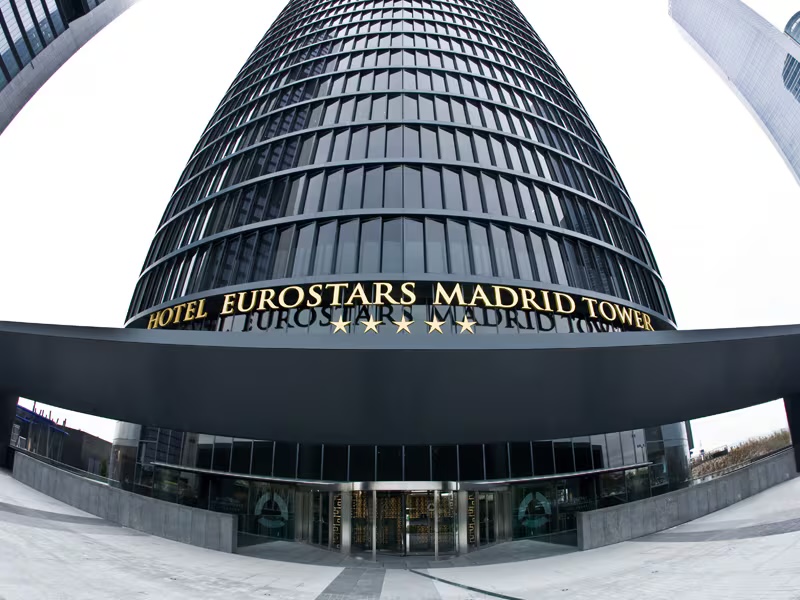 Looking up the curved glass and steel tower from the forecourt, with the gold 'Hotel Eurostars Madrid Tower' lettering above the entrance canopy and revolving doors.