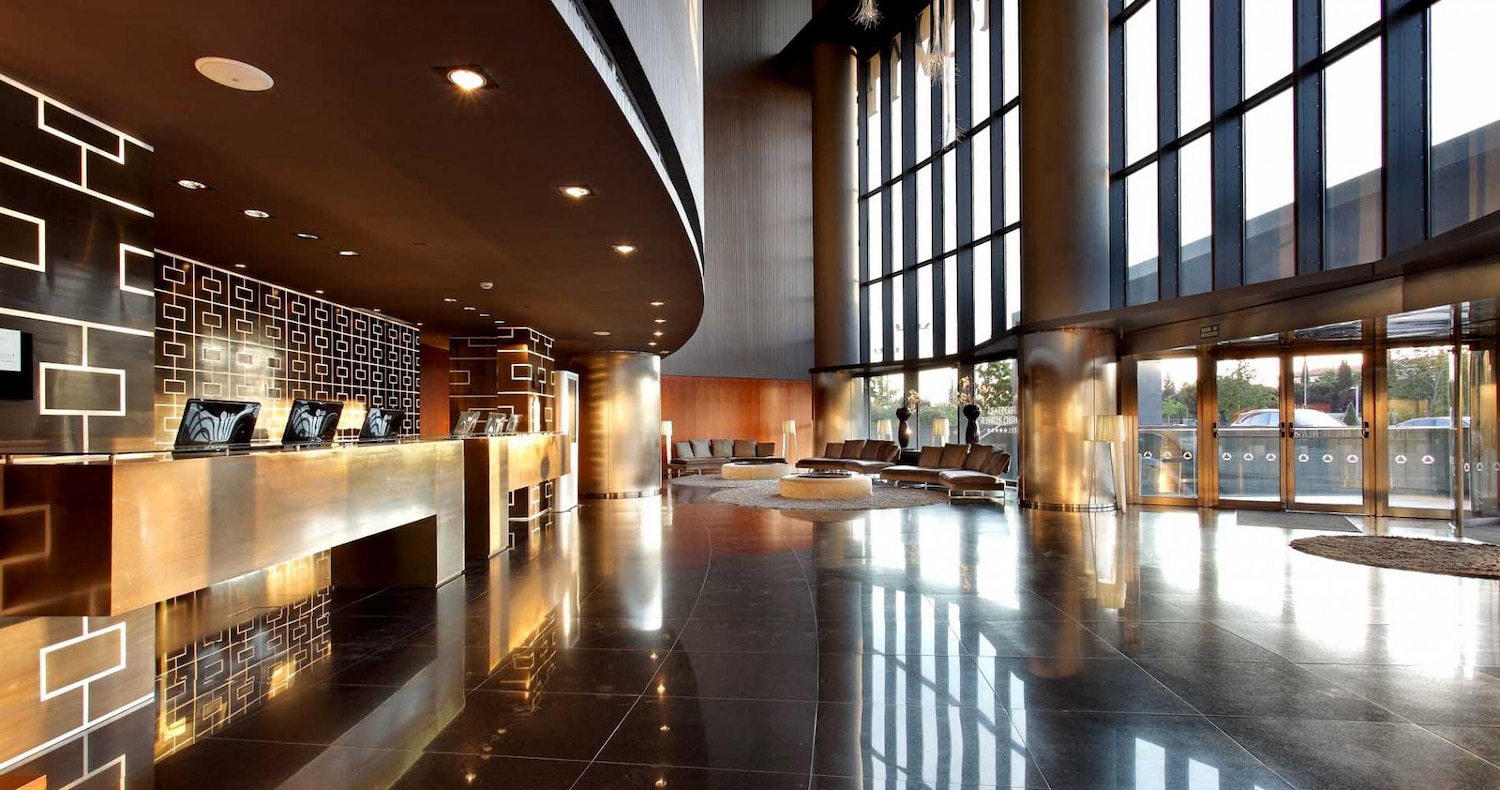 The double-height hotel lobby with a long metal reception desk, patterned dark wall panels, a polished black stone floor and floor-to-ceiling windows beside the entrance doors.