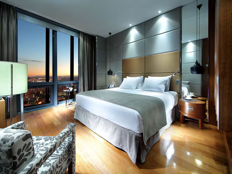 A guest room with a wide bed in white and grey linen, a padded headboard wall, black pendant lamps, a patterned armchair and a floor-to-ceiling window over the city lights at dusk.
