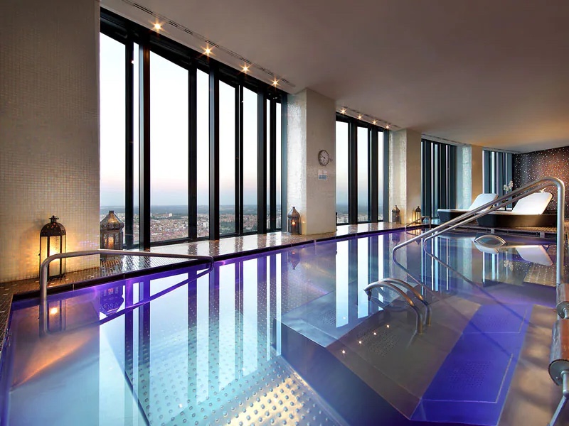 An indoor spa pool with steel handrails and lanterns along its edge, floor-to-ceiling windows looking out over the city at dusk, and two loungers at the far end.