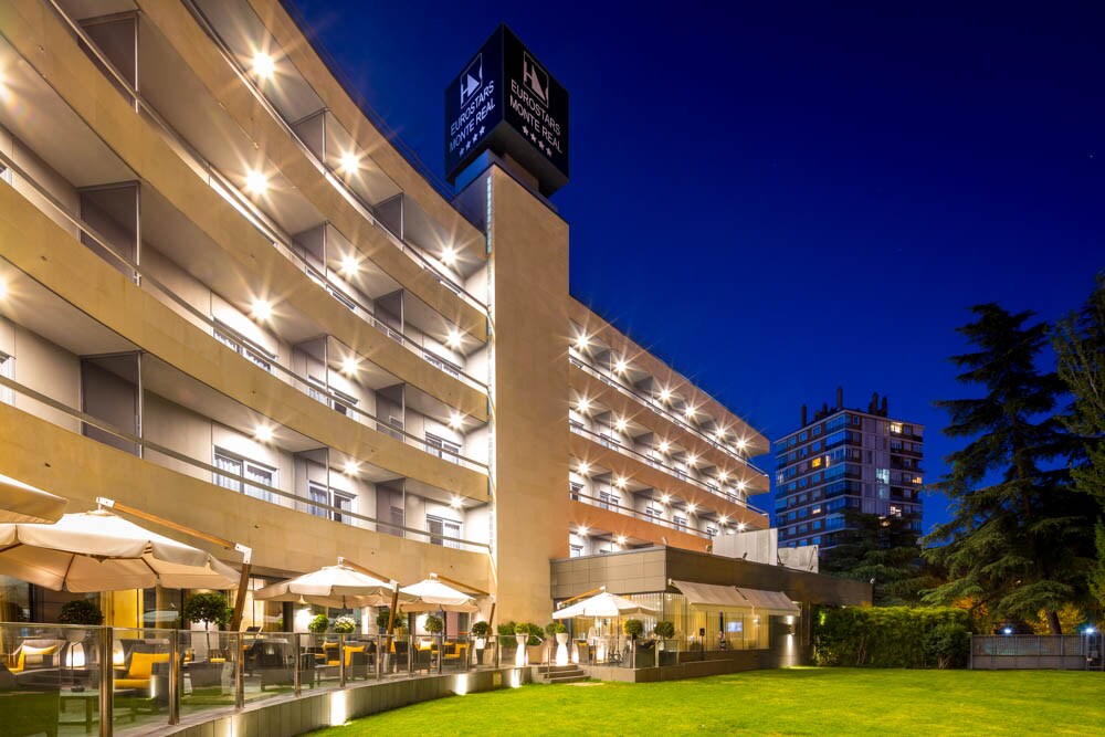 The hotel's facade lit up at night, with balconies, a garden terrace with umbrellas, and the Eurostars Monte Real sign on top.