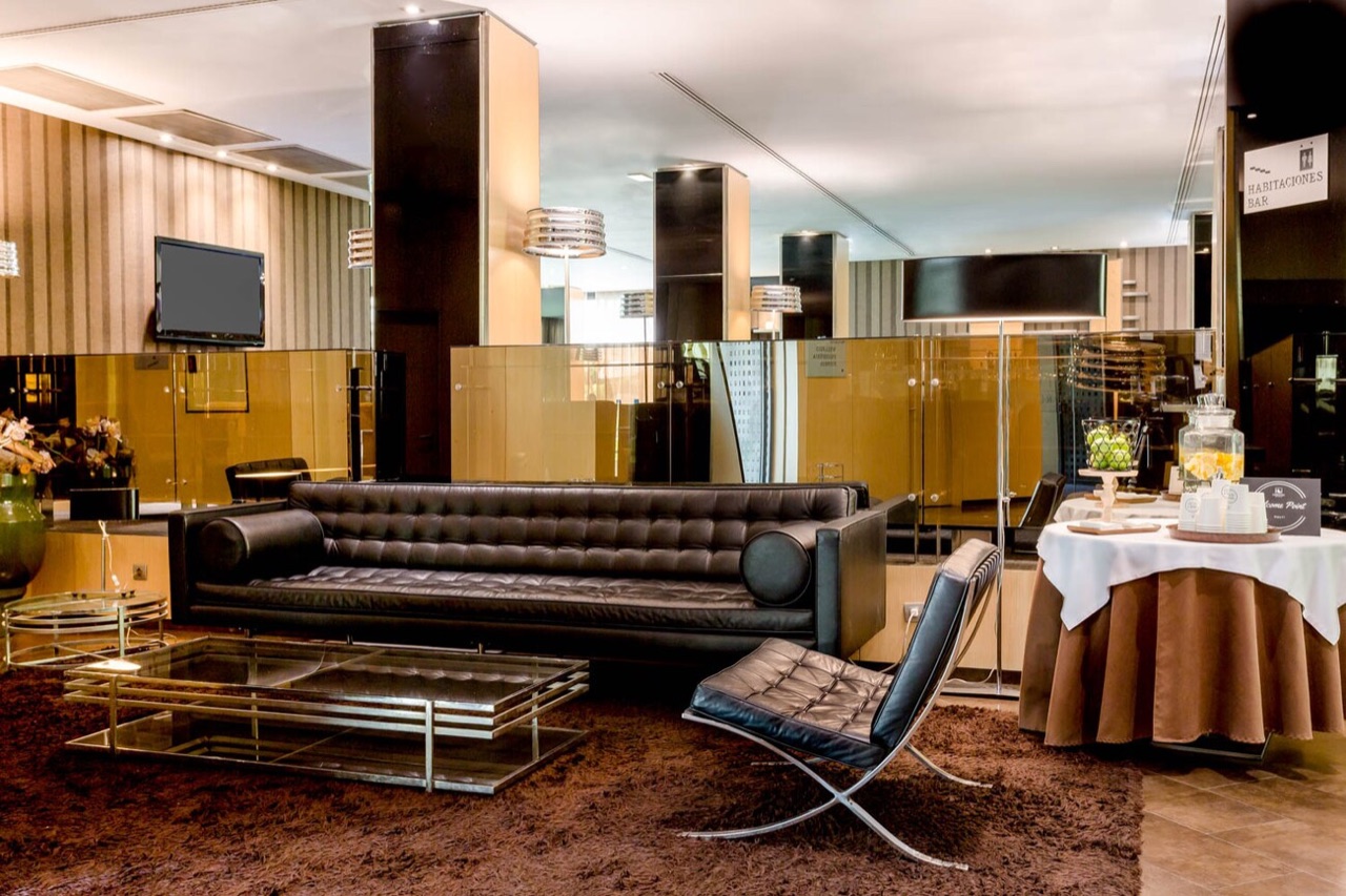 A lobby seating area with a dark leather sofa and armchair, a glass coffee table, and a mirrored wall with a wall-mounted TV.