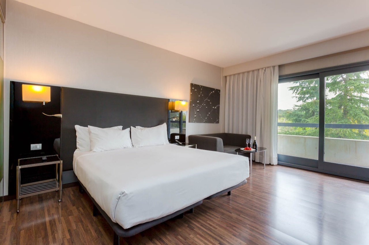 A hotel room with a white king bed, a grey armchair, and a glass sliding door open to a private terrace with trees beyond.