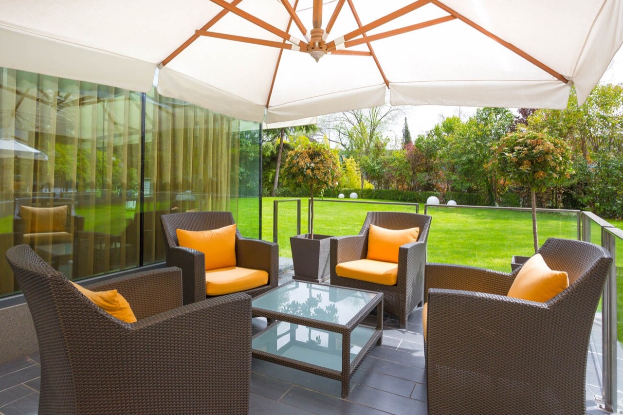 An outdoor terrace with wicker armchairs and orange cushions under a large umbrella, facing the hotel's lawn.