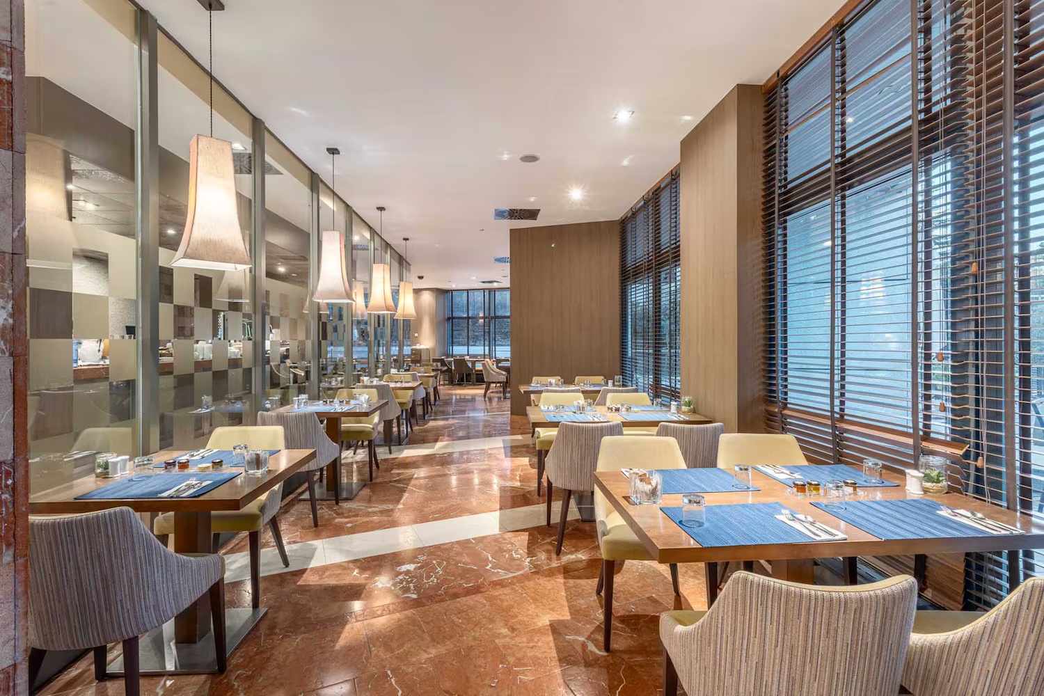 The hotel restaurant with square wooden tables laid with blue place mats and condiments, cream and striped chairs, red marble floor, pendant lamps and slatted blinds on tall windows.