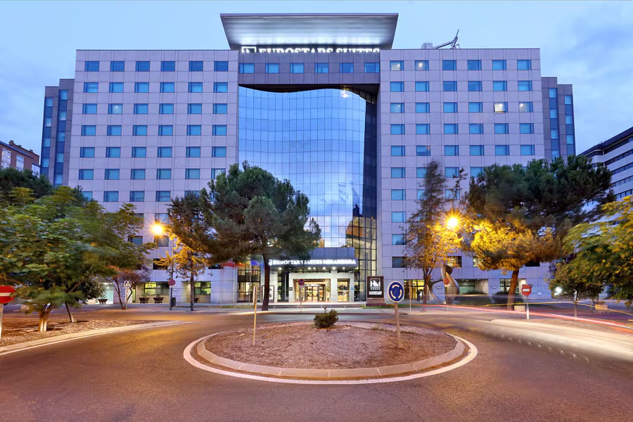 The hotel's white-panelled facade with a curved blue-glass central bay, seen across a roundabout with pine trees and street lamps at dusk.