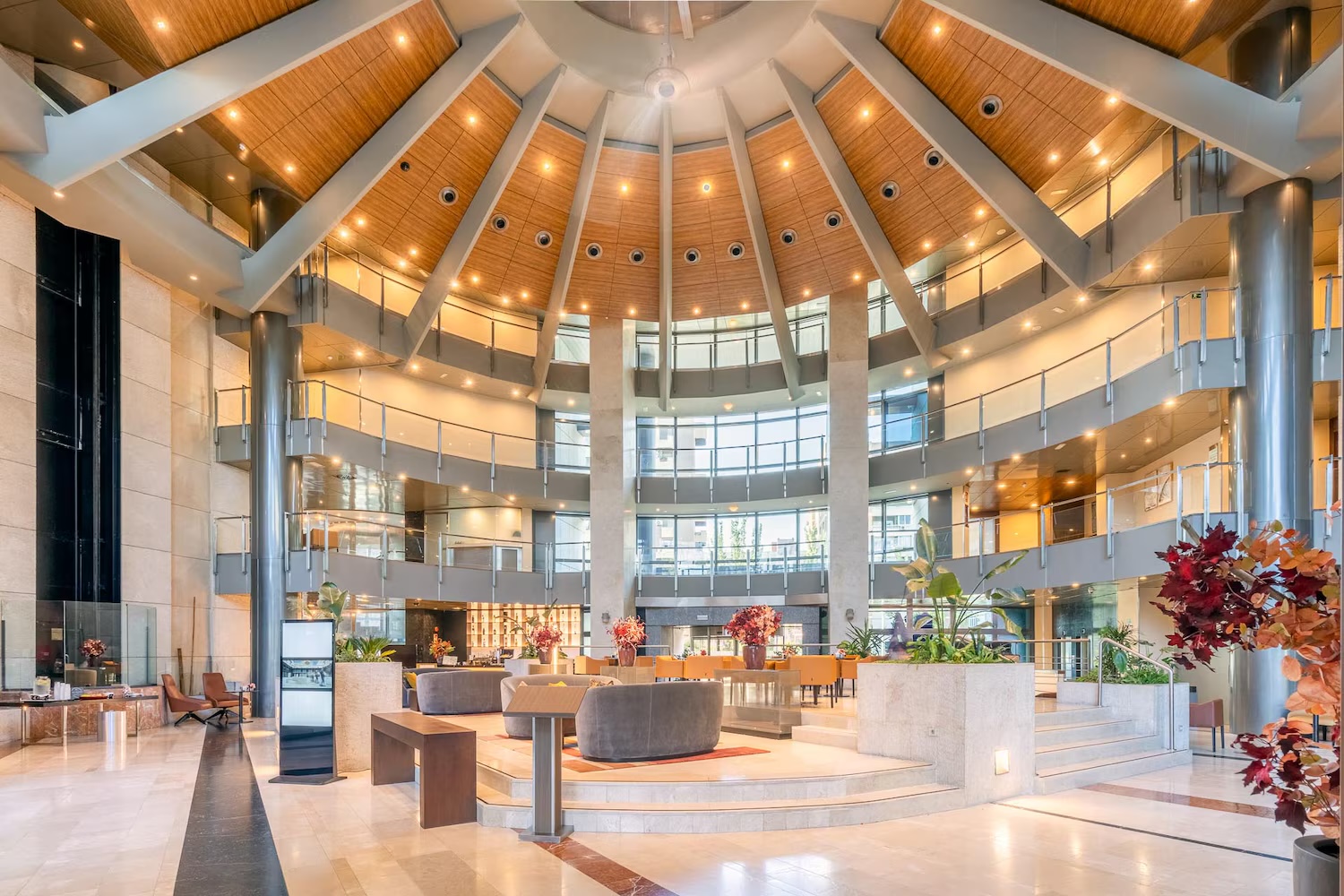A multi-storey atrium lobby under a radial wooden ceiling, with curved balconies on each floor, grey sofas and a table on a raised platform, and planters with red-leafed shrubs.