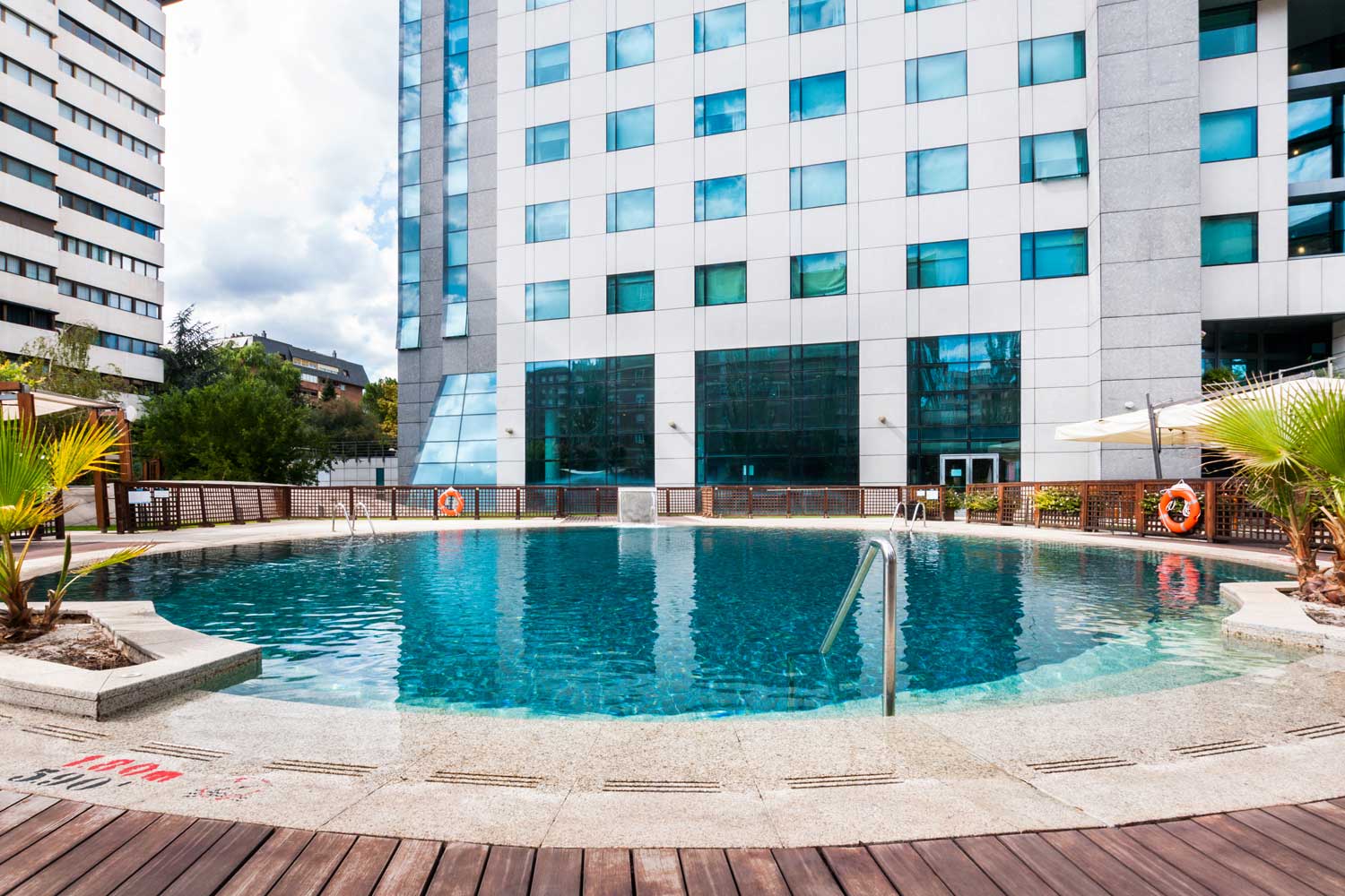 A curved outdoor pool with a stainless-steel ladder, set in a stone and wood deck ringed by palm plants and a lattice fence, with the hotel's white-panelled glass facade rising behind.