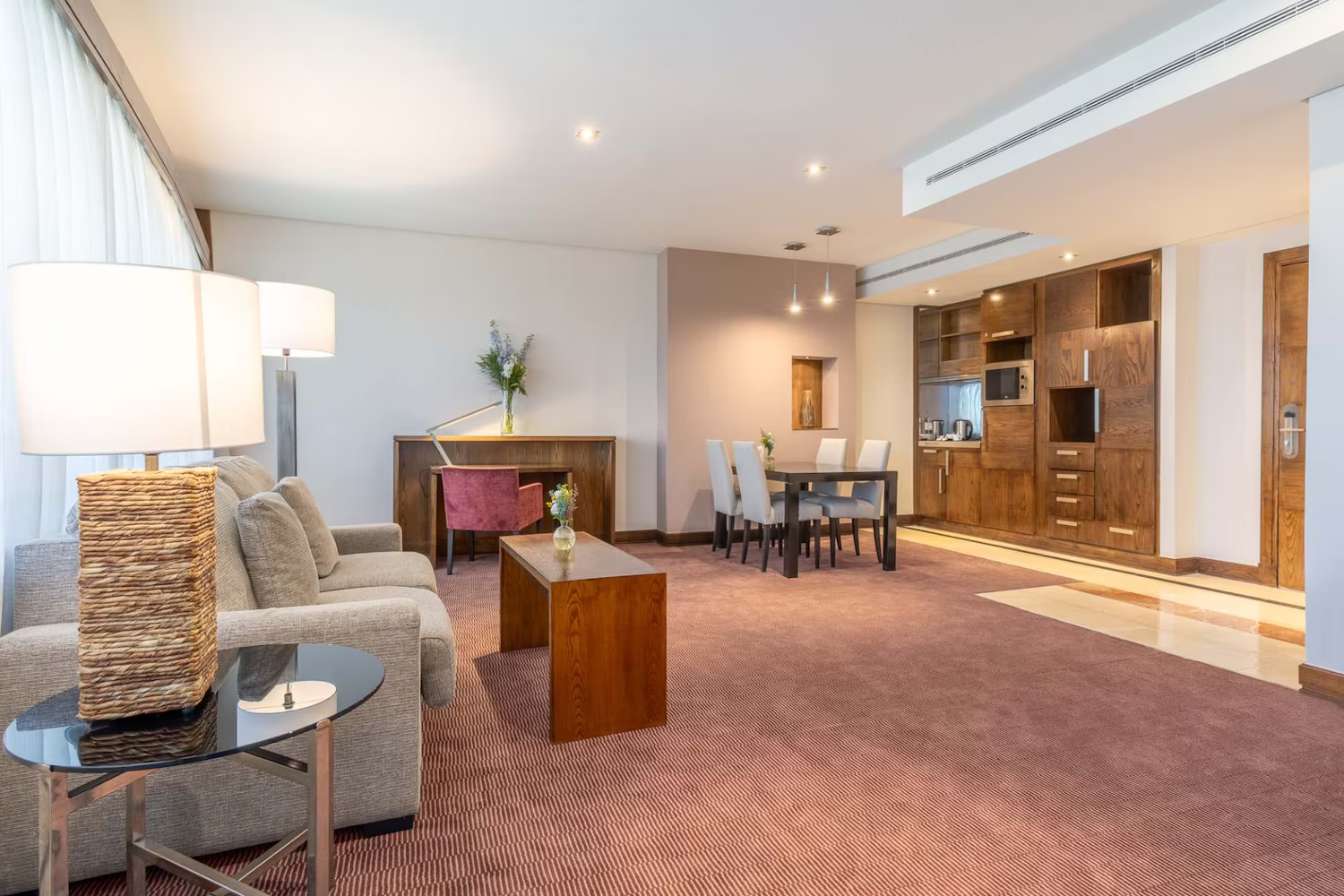 A suite living room with a beige sofa and wicker-based lamps, a wooden desk with a red chair, a four-seat dining table and a wooden kitchenette wall with a microwave and cabinets.