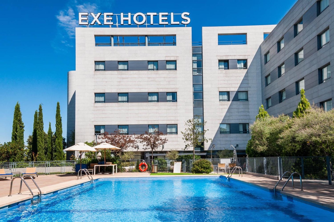 The hotel's outdoor swimming pool with sun loungers and umbrellas set against the building, topped with an EXE HOTELS sign.