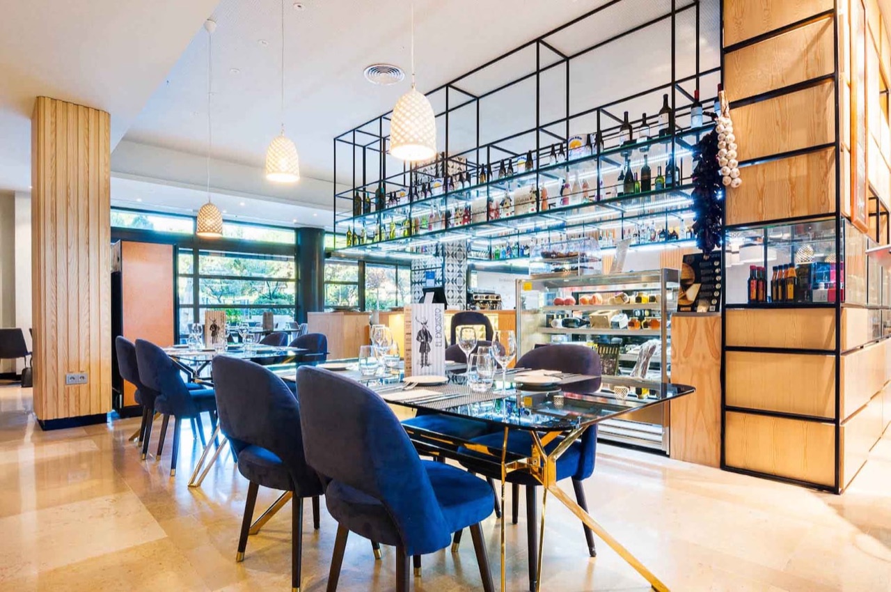 The El Exquisito dining room with blue velvet chairs at set tables in front of a lit shelf of wine bottles and a deli counter.