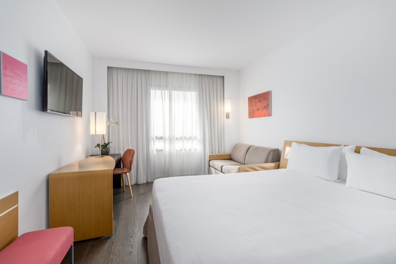 A guest room with a white bed, a grey sofa by the curtained window, a wall-mounted TV and a wooden desk.
