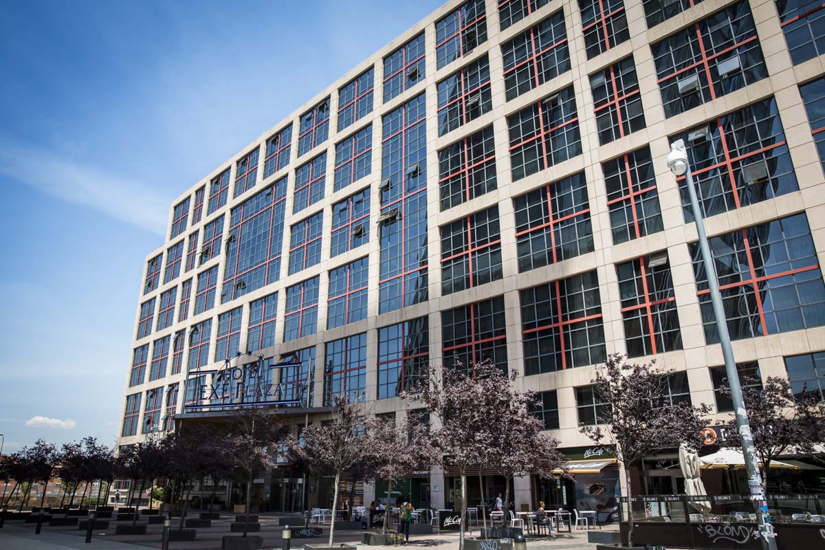 The Exe Plaza hotel's glass and stone facade under a blue sky, with the hotel's sign above the entrance and trees and a McCafe storefront at street level.