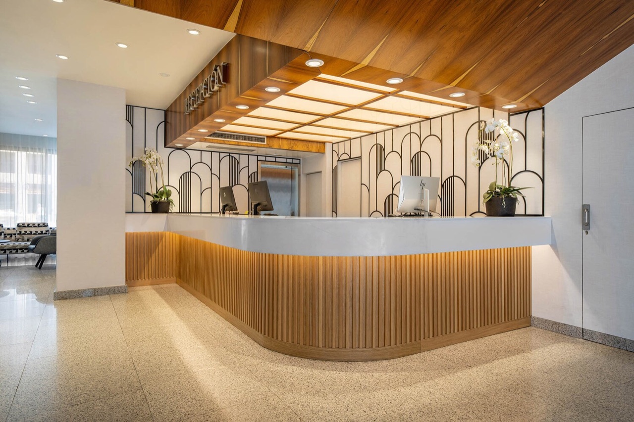 A curved wood-paneled reception desk with two computer monitors, orchid plants, and an Art Deco-style metal screen on the wall behind it, under a wood-slat ceiling.