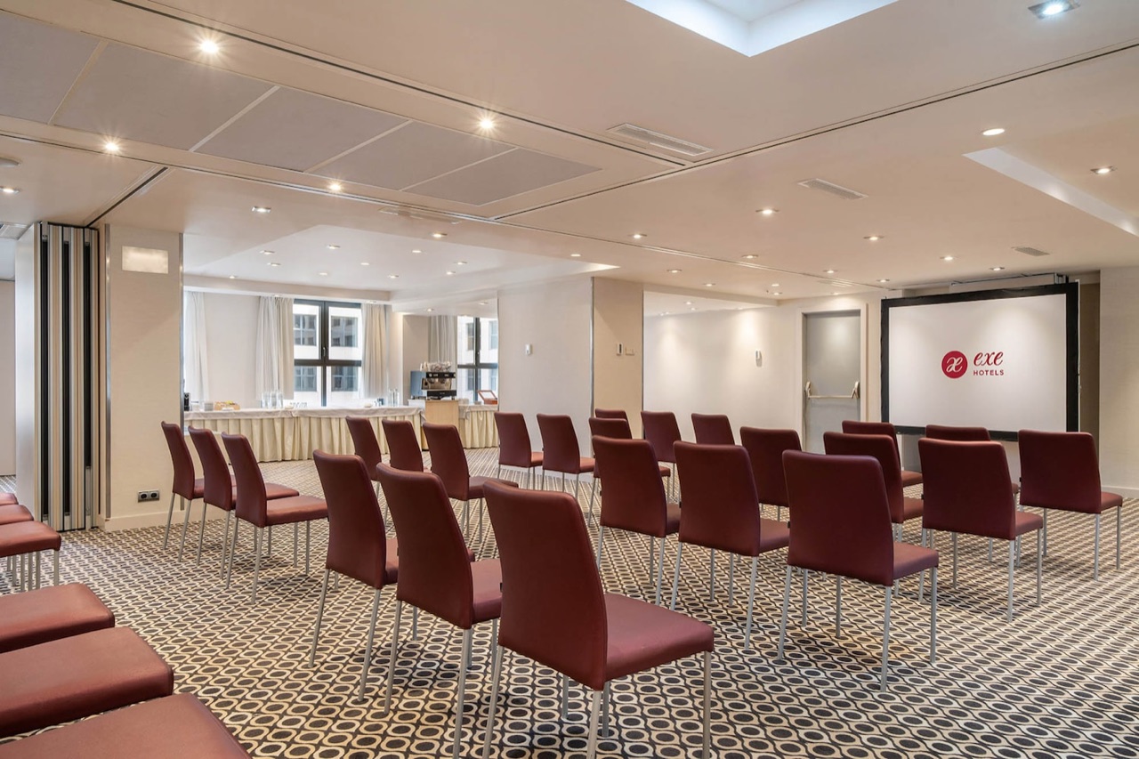 A meeting room set theatre-style with rows of burgundy chairs facing a projector screen displaying the Exe Hotels logo, with a coffee station and windows to one side.