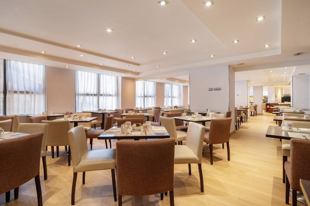 A restaurant dining room set for breakfast with rows of tables and upholstered chairs, large curtained windows along one wall, and a buffet counter visible in the back room.
