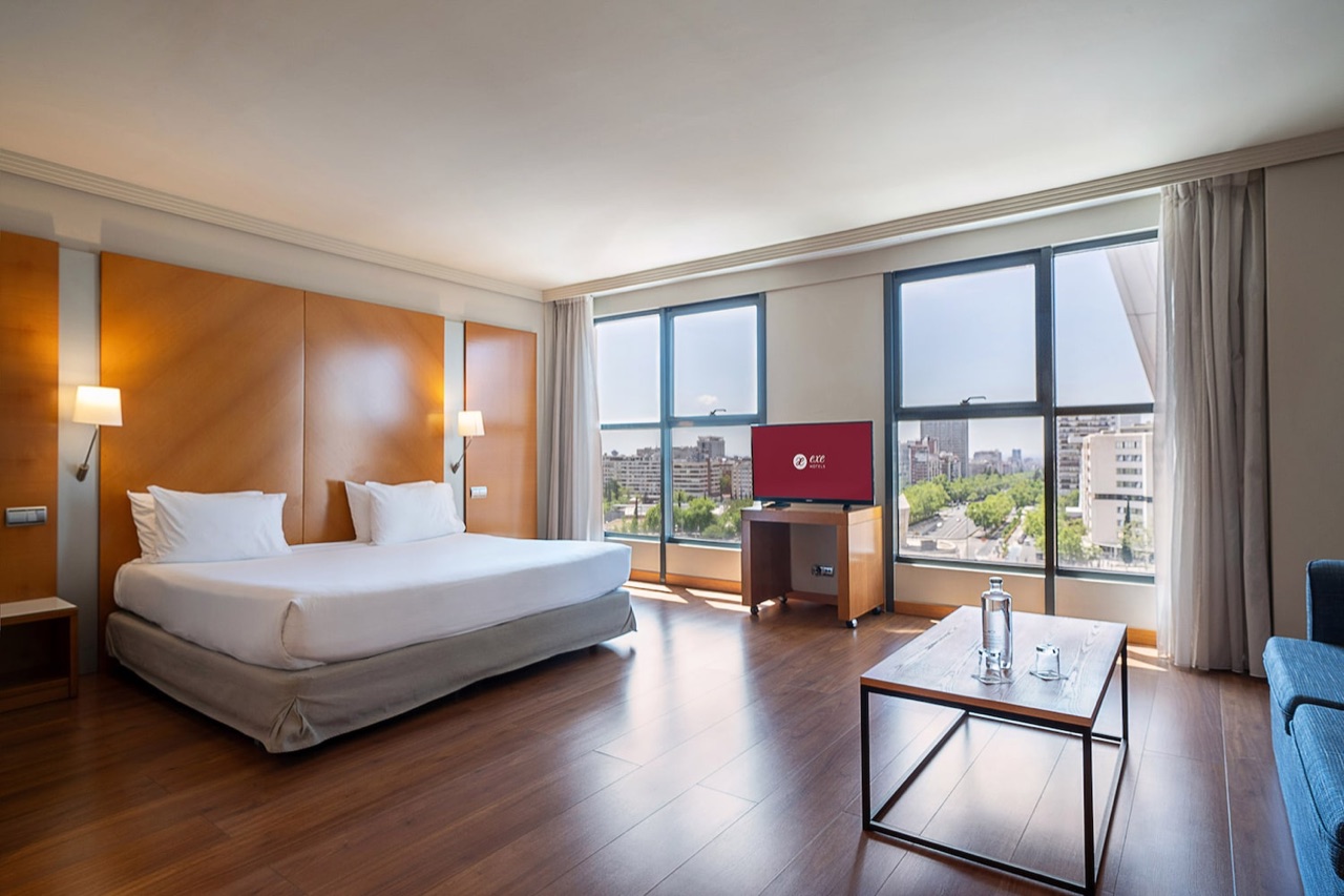A hotel room with a bed against a wood-paneled headboard, a television on a rolling cart displaying the Exe Hotels logo, a small coffee table, and floor-to-ceiling windows overlooking the city.