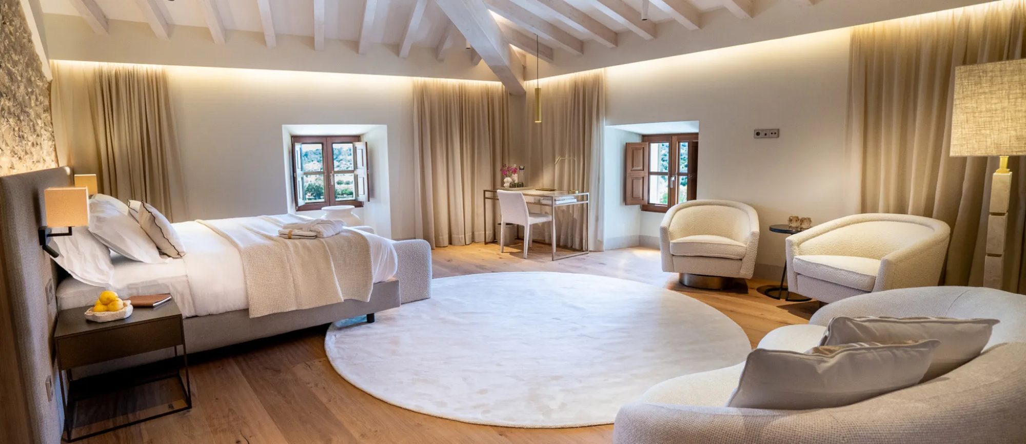 Suite at Finca Banyols with a white beamed ceiling, a king bed, a round white rug and cream armchairs beside wood-shuttered windows