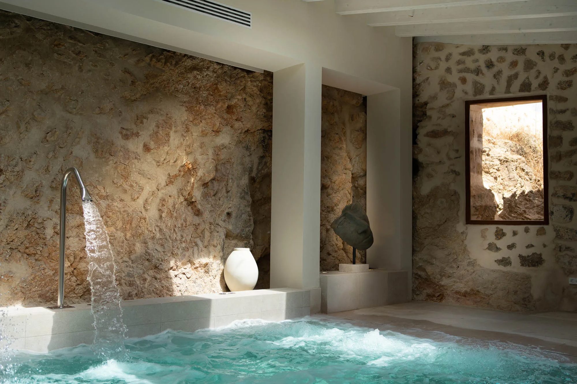 Indoor spa pool at Finca Banyols set against a rustic exposed stone wall, with a wall-mounted water spout and a ceramic urn beside a sculpture