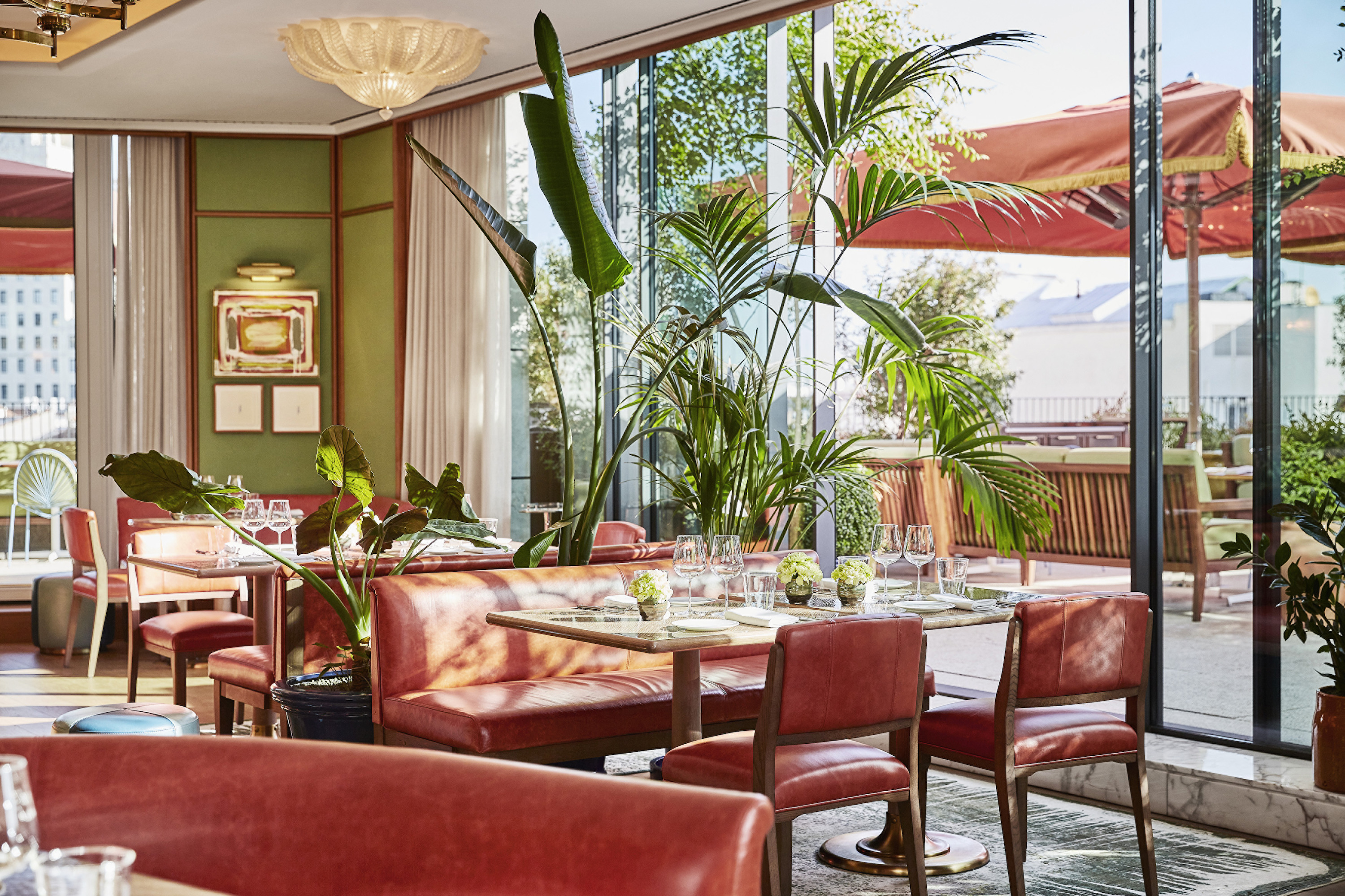 Restaurant dining room with coral leather banquettes, marble-topped tables, tall plants and glass walls onto a terrace with red parasols.