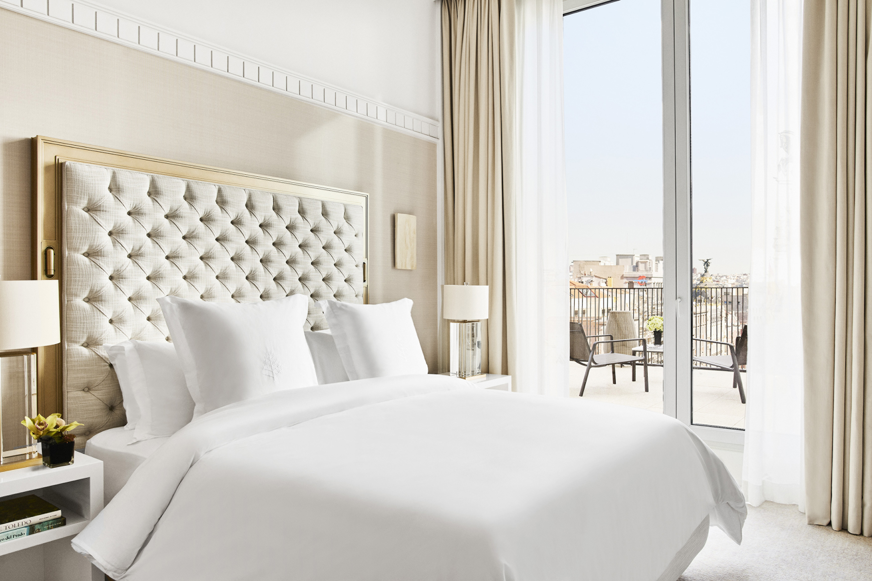 Guest room with a tufted headboard, white bedding and glass doors opening onto a furnished terrace overlooking city rooftops.