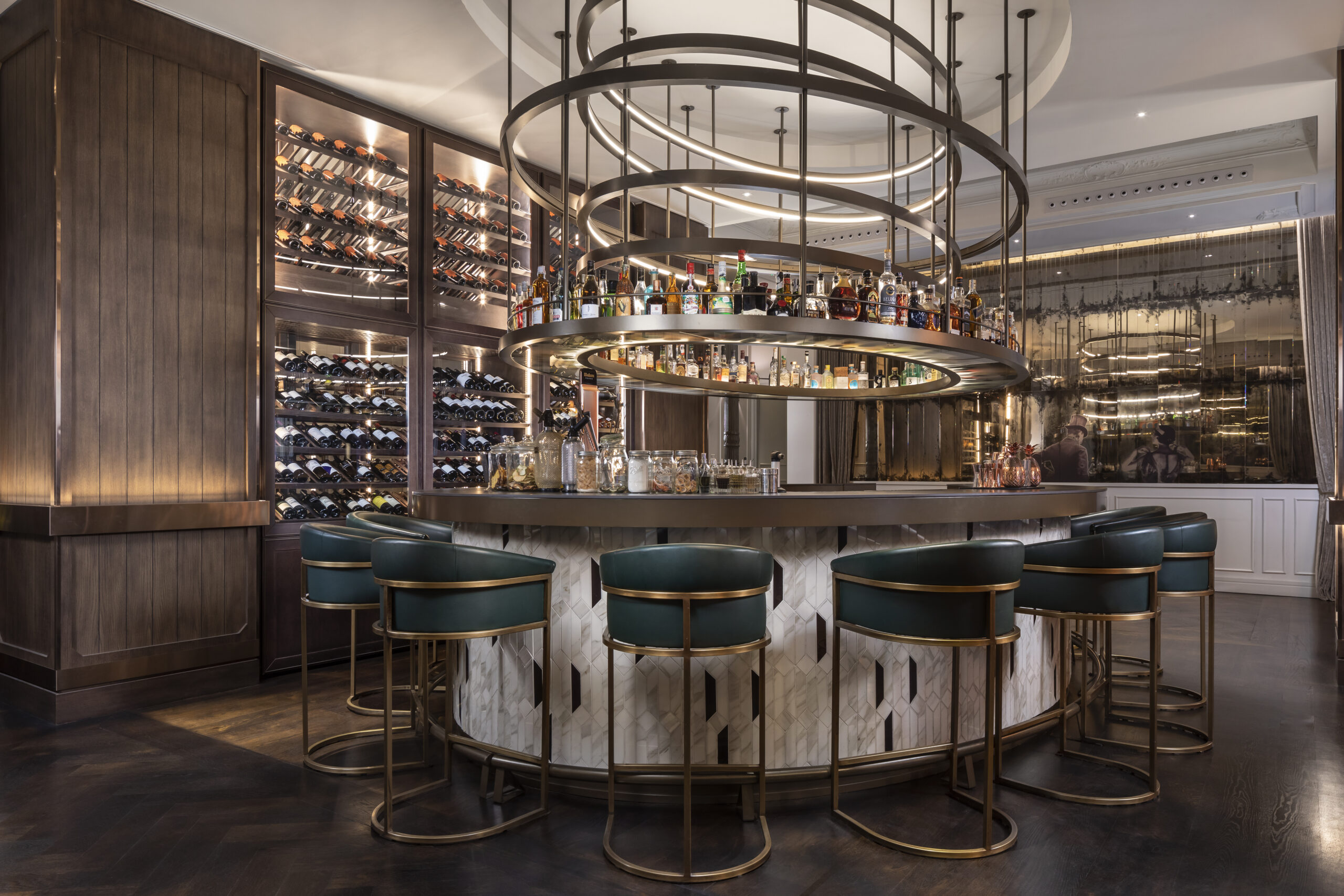 A circular bar with a white and black tiled front, green leather stools on brass frames, a spiral ring chandelier holding bottles above the counter, and a backlit wall of wine racks.