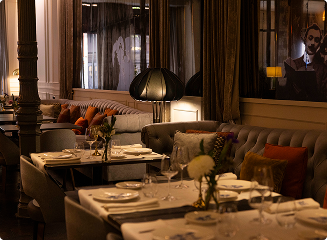 A dining lounge with tables set with glassware and flowers beside grey sofas with orange cushions, a dark standing lamp and a framed portrait on the wall.