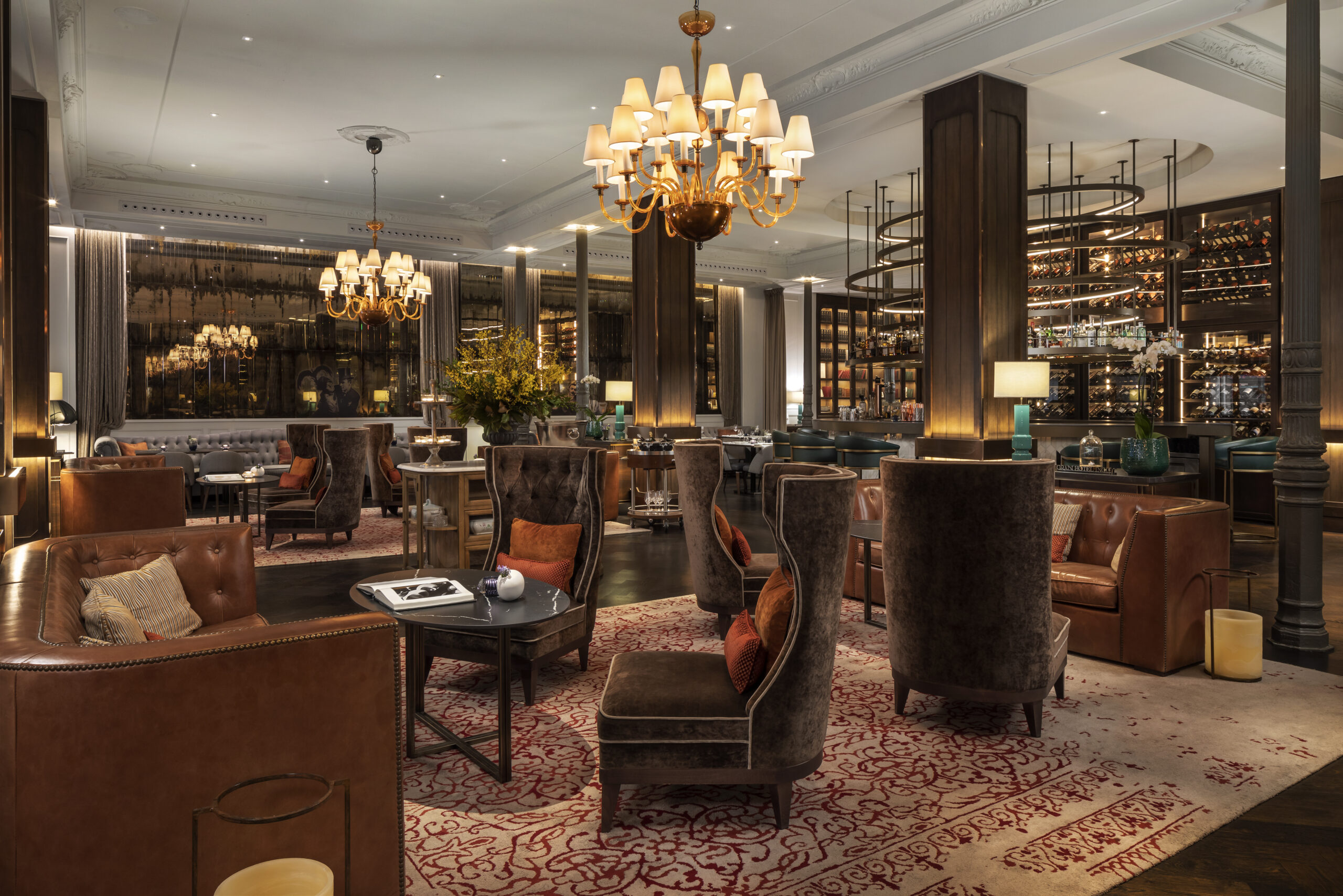 A lobby lounge with tall brown velvet wing chairs and tan leather sofas on a red-patterned rug, brass chandeliers with cream shades, and a bar with a backlit wine wall behind.
