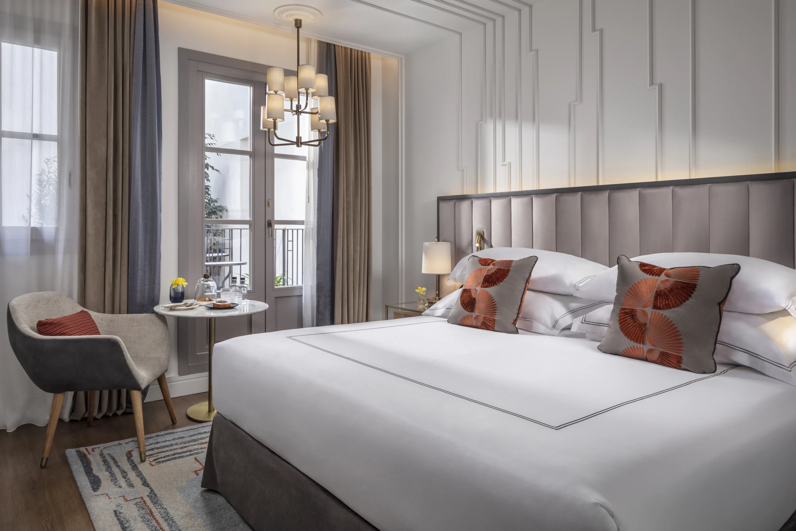 A guest room with a wide bed under a tall grey padded headboard, orange fan-patterned cushions, a small marble side table with a grey armchair, and a French window with balcony railings and beige curtains.