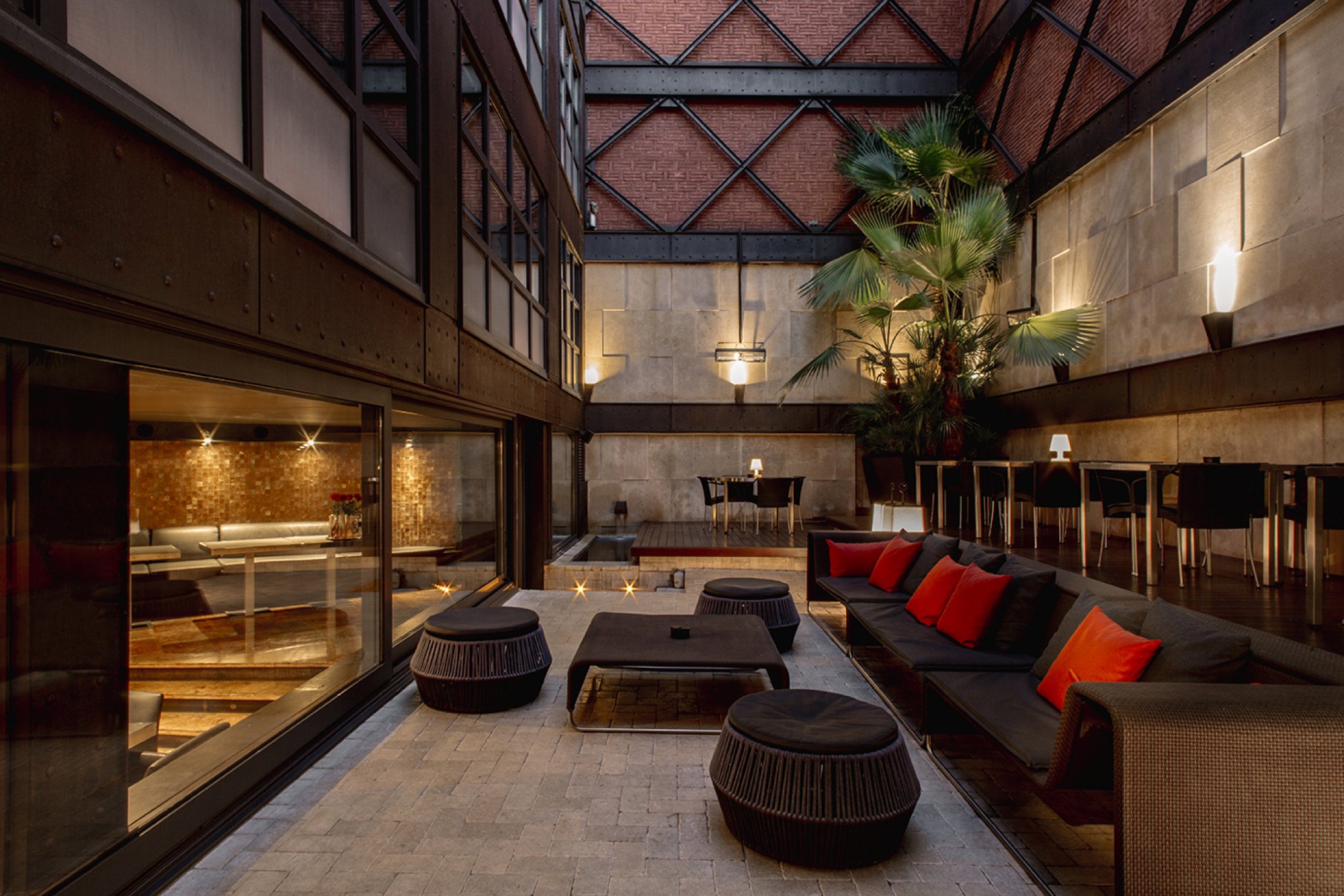 Lobby atrium at Hotel Granados 83 under a steel-and-glass roof, with red-brick walls, dark sofas and red cushions