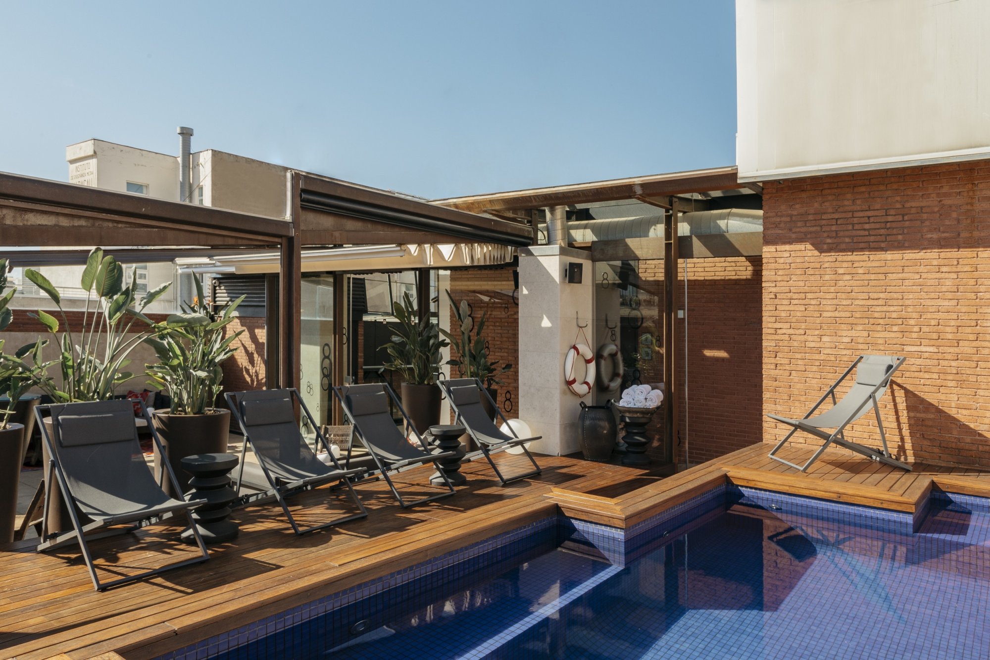 Top-floor terrace at Hotel Granados 83 with wooden decking, black sun loungers beside the pool and an exposed-brick wall