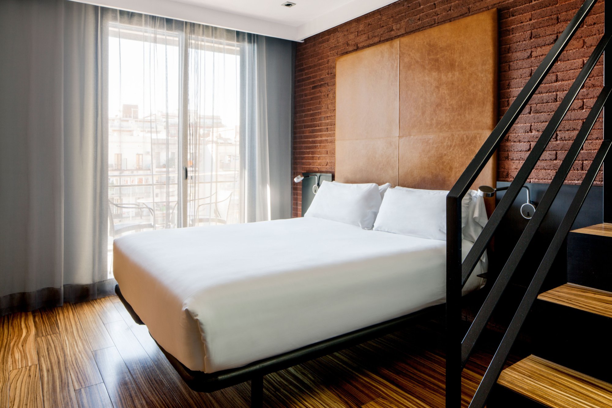 Guest room at Hotel Granados 83 with a white bed, exposed-brick walls, wooden floors and a black metal staircase to an upper level